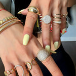 Close-up of two hands wearing multiple gold rings with pavé crystals and bold designs, paired with gold bracelets, styled with pastel yellow manicure.