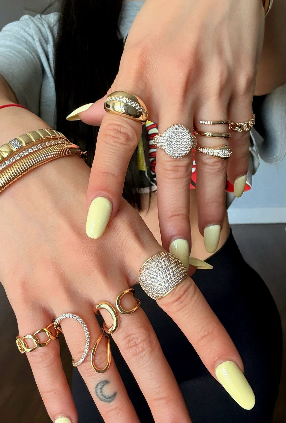 Close-up of two hands wearing multiple gold rings with pavé crystals and bold designs, paired with gold bracelets, styled with pastel yellow manicure.