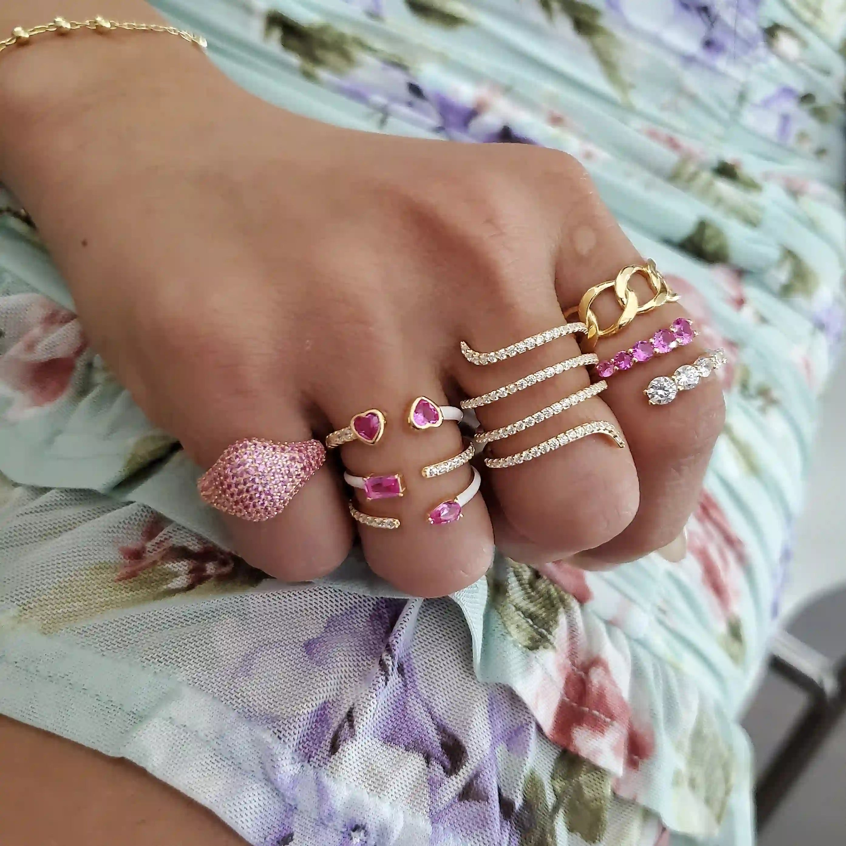 Close-up of a hand wearing multiple gold rings with pink gemstones and clear crystals, styled with a floral print outfit.