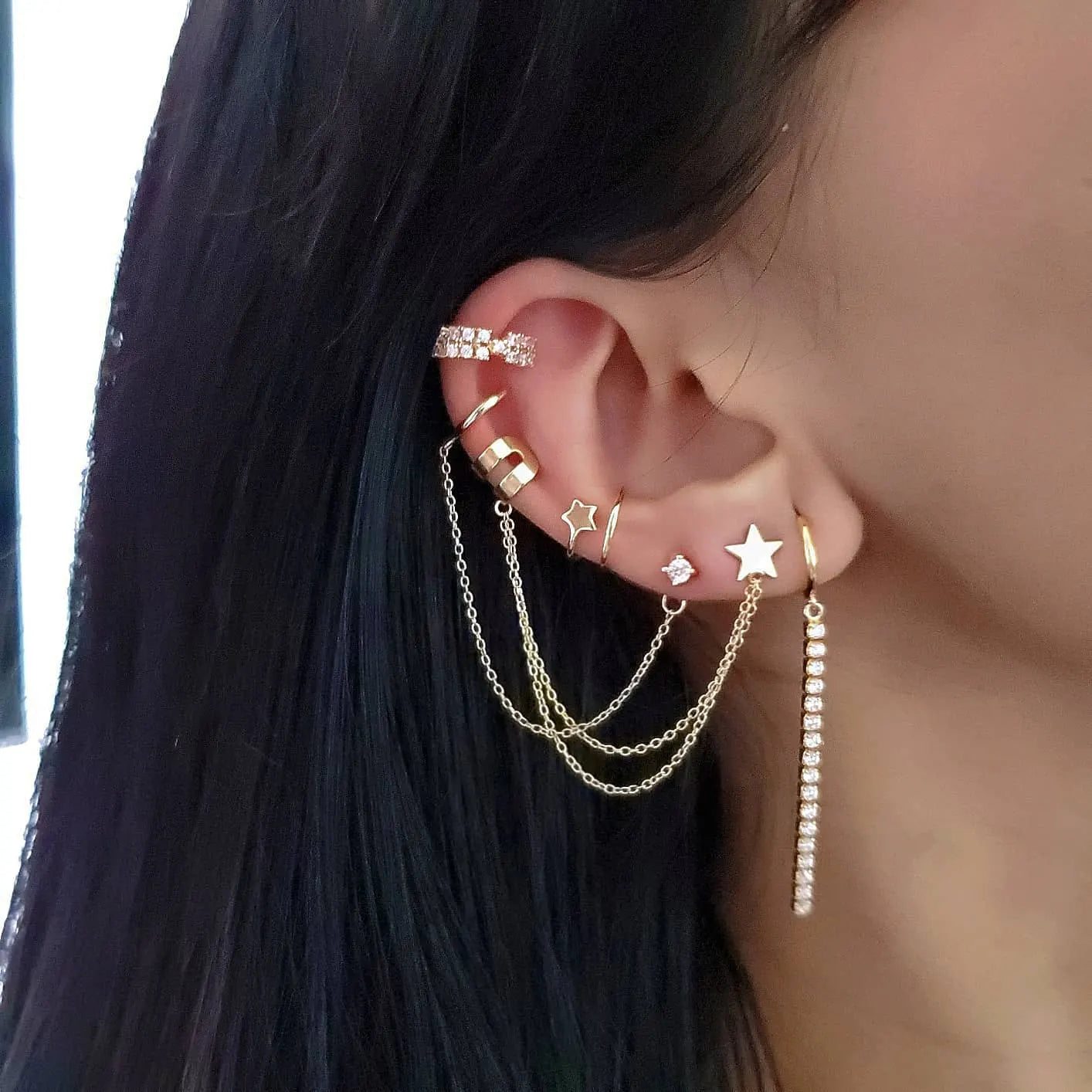 Close-up of an ear adorned with multiple gold earrings, including hoops, star-shaped studs, chain details, and a long rhinestone drop earring.