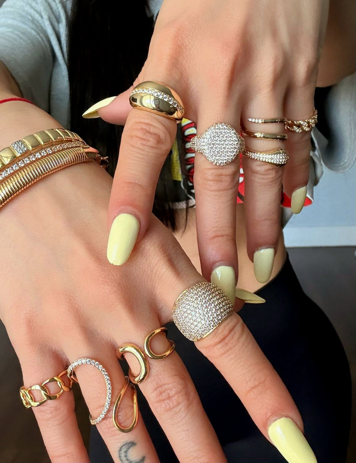 Close-up of two hands wearing multiple gold rings with pavé crystals and bold designs, paired with gold bracelets, styled with pastel yellow manicure.
