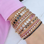 Close-up of a woman's wrist stacked with multiple gold bracelets featuring heart-shaped charms and rectangular stones in black, pink, and clear designs, paired with sparkling rhinestone accents.