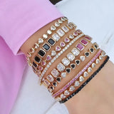 Close-up of a woman's wrist stacked with multiple gold bracelets featuring heart-shaped charms and rectangular stones in black, pink, and clear designs, paired with sparkling rhinestone accents.